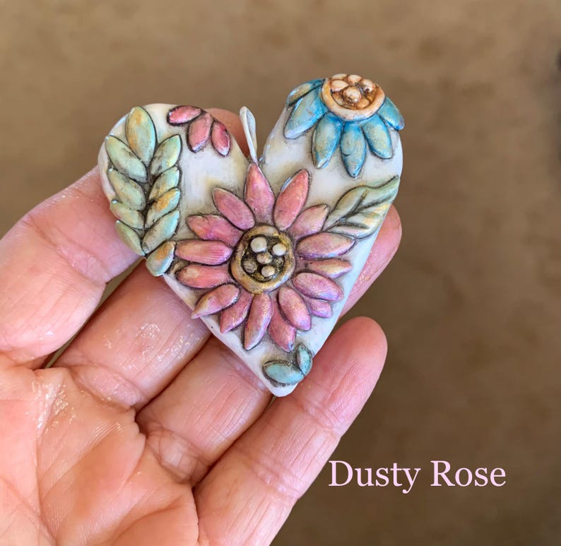 Puede incluir: Un adorno de cer&aacute;mica en forma de coraz&oacute;n con fondo blanco y dise&ntilde;os florales. Las flores son rosas, azules y verdes con detalles dorados. El texto "Dusty Rose" est&aacute; escrito debajo del adorno.