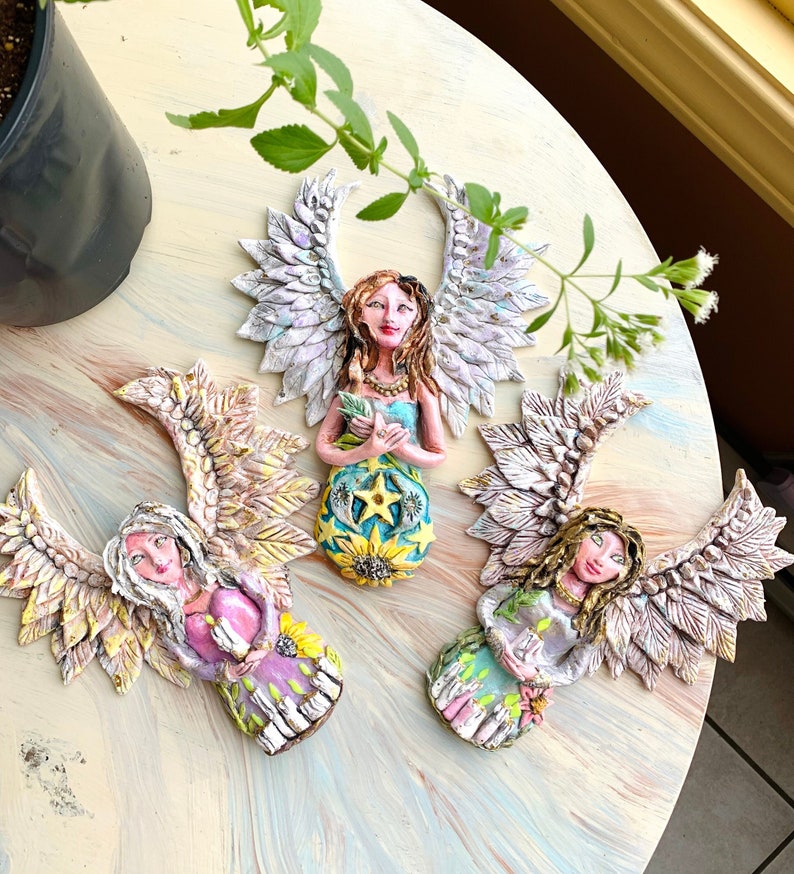 May include: Three handmade ceramic angels with wings, each holding a small object. The angels are painted in pastel colors with floral details and have a whimsical, fairy-like appearance.