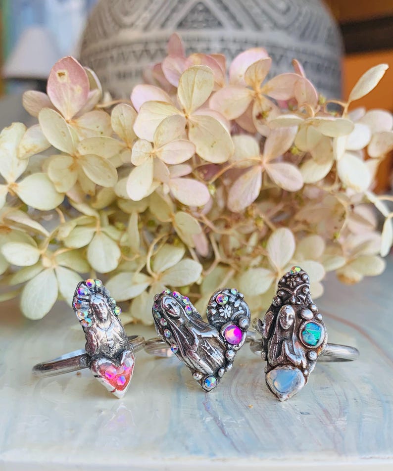 May include: Three silver rings with intricate designs and iridescent details. Each ring showcases a unique sculpted figure and colourful gemstones. The rings are presented against a backdrop of dried, pale-coloured flowers.