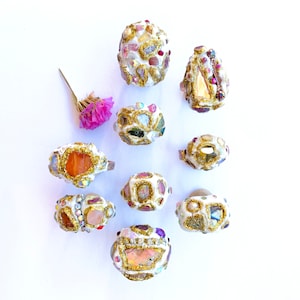 May include: Ten unique rings with a variety of colorful stones and gold accents. The rings are set on a white background.