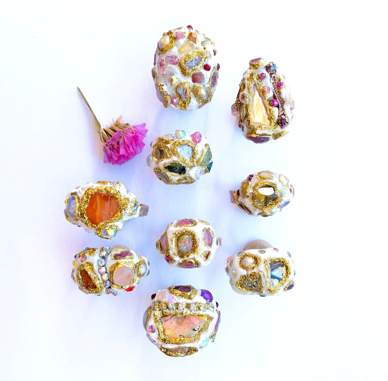 May include: Ten unique rings with a variety of colorful stones and gold accents. The rings are set on a white background.