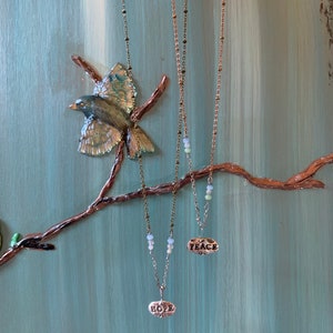 Puede incluir: Dos collares de oro con cadenas delicadas y peque&ntilde;os colgantes ovalados. Los colgantes son de plata y tienen las palabras "HOPE" y "PEACE" inscritas en ellos. Los collares cuelgan de una rama sobre un fondo azul claro.