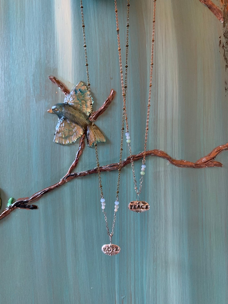 Puede incluir: Dos collares de oro con cadenas delicadas y peque&ntilde;os colgantes ovalados. Los colgantes son de plata y tienen las palabras "HOPE" y "PEACE" inscritas en ellos. Los collares cuelgan de una rama sobre un fondo azul claro.