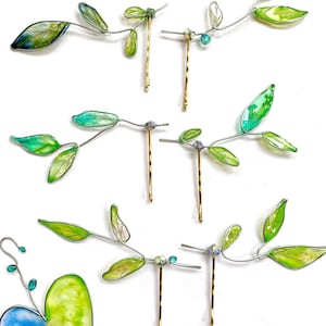 May include: Hair accessories featuring delicate leaf designs. The leaves are a mix of green and yellow, with a touch of blue, attached to silver wire. The bobby pins are gold-colored, and some have small, sparkling embellishments.