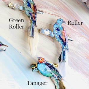 May include: Three hair clips featuring bird designs labeled Green Roller, Roller, and Tanager. Each clip has a metallic base, iridescent crystals, and a long feather tail. The birds are decorated with blue, green, and orange colors and glitter.