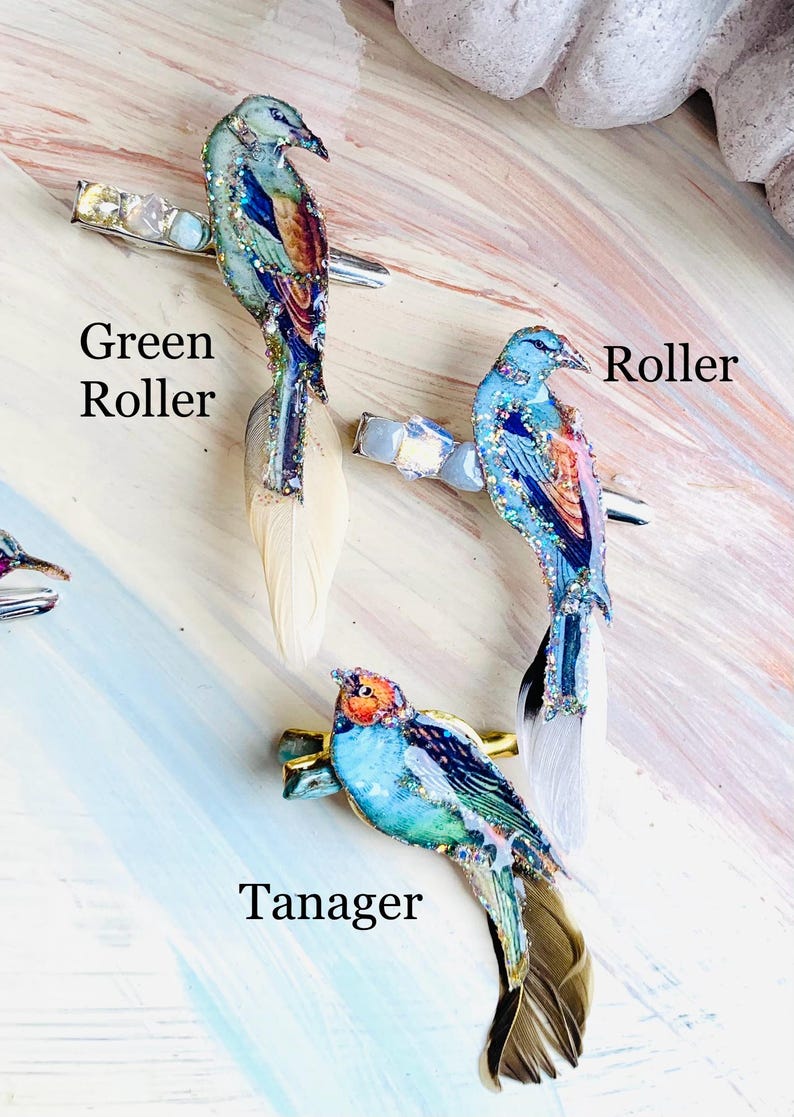 May include: Three hair clips featuring bird designs labeled Green Roller, Roller, and Tanager. Each clip has a metallic base, iridescent crystals, and a long feather tail. The birds are decorated with blue, green, and orange colors and glitter.