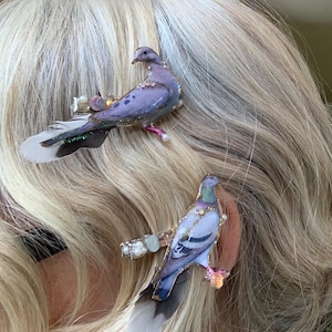 May include: Two hair clips featuring detailed pigeon figures in shades of purple, grey, and blue. Each bird is accented with small white pearls and iridescent glitter, attached to a metal clip base with soft, natural grey and white feathers.