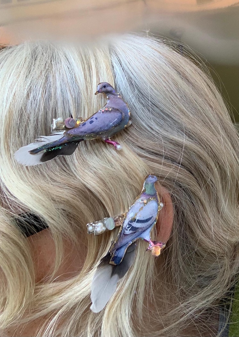 May include: Two hair clips featuring detailed pigeon figures in shades of purple, grey, and blue. Each bird is accented with small white pearls and iridescent glitter, attached to a metal clip base with soft, natural grey and white feathers.