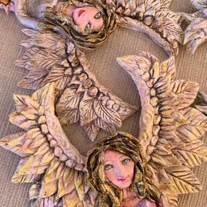 May include: Two ceramic angels with wings made of leaves. The angels are painted in shades of white, gold, and pink. They are holding flowers in their hands.