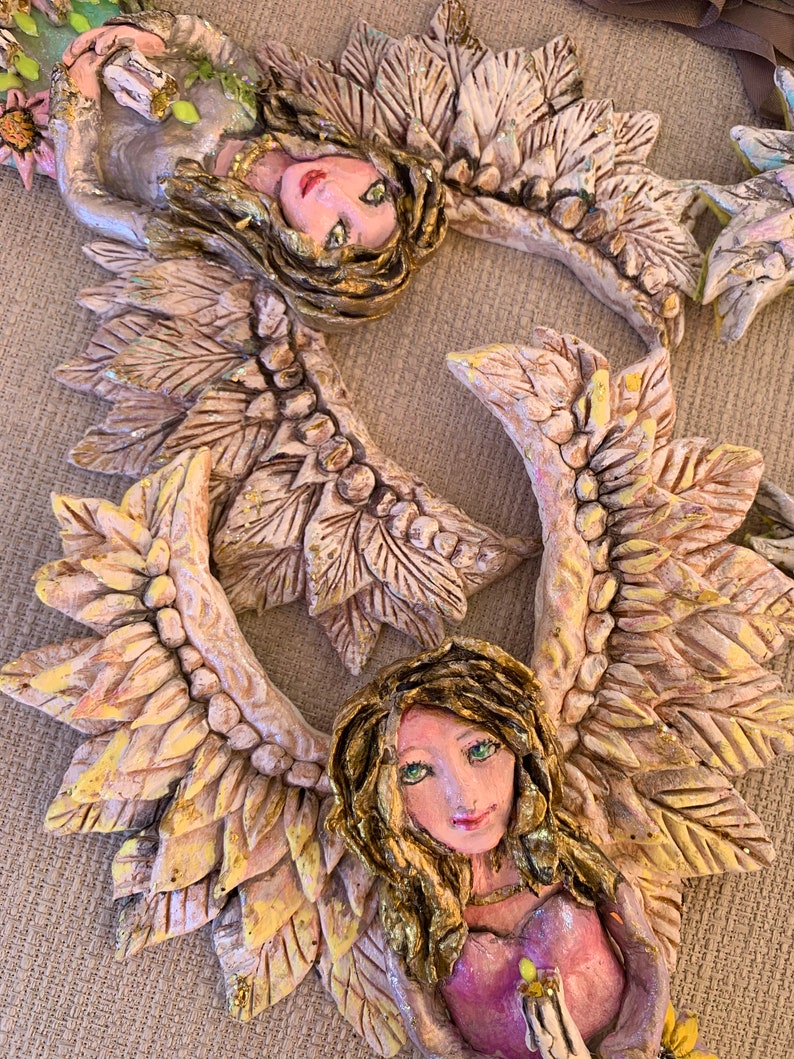 May include: Two ceramic angels with wings made of leaves. The angels are painted in shades of white, gold, and pink. They are holding flowers in their hands.