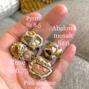 May include: Five handmade rings with different gemstone inlays. The rings are made of white clay with gold accents. The stones include rose quartz, abalone, pyrite, and pink abalone. The rings are labeled with their names and sizes.