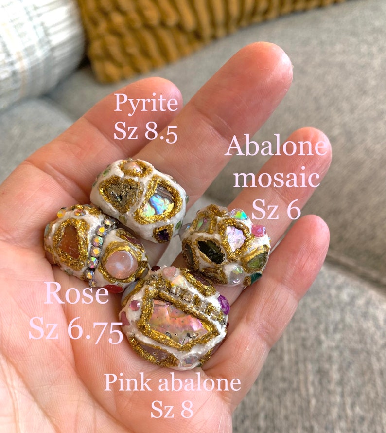 May include: Five handmade rings with different gemstone inlays. The rings are made of white clay with gold accents. The stones include rose quartz, abalone, pyrite, and pink abalone. The rings are labeled with their names and sizes.