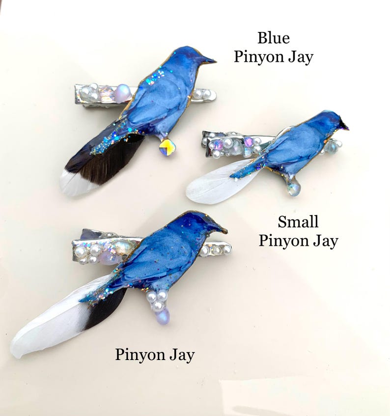 May include: Three hair clips featuring bird designs labeled Blue Pinyon Jay, Small Pinyon Jay, and Pinyon Jay. Each clip displays a blue bird figure adorned with white pearls, iridescent beads, and feathers in shades of white, black, and blue.