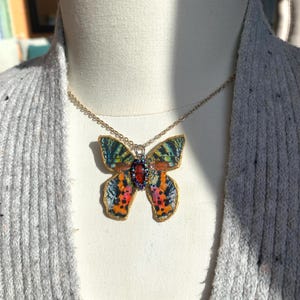 Puede incluir: Un collar de cadena dorada con un colgante de mariposa colorido. La mariposa tiene un patr&oacute;n rojo, naranja, amarillo, verde y azul. El colgante tiene una peque&ntilde;a piedra preciosa roja en el centro.