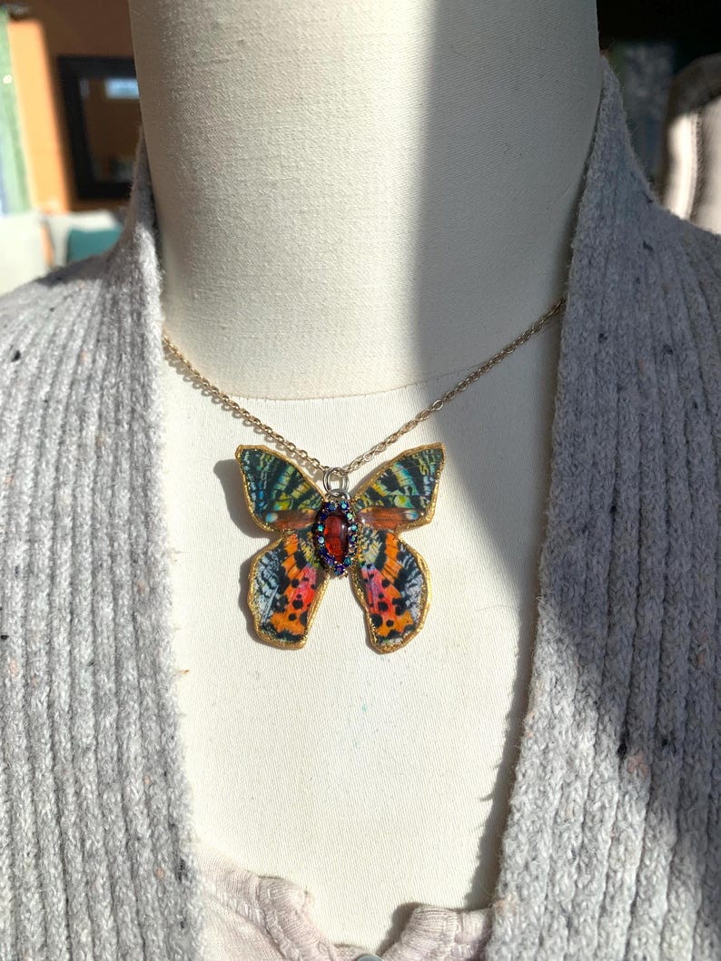 Puede incluir: Un collar de cadena dorada con un colgante de mariposa colorido. La mariposa tiene un patr&oacute;n rojo, naranja, amarillo, verde y azul. El colgante tiene una peque&ntilde;a piedra preciosa roja en el centro.