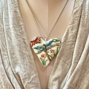 Puede incluir: Un collar con un colgante de coraz&oacute;n de cer&aacute;mica blanca y una cadena plateada. El coraz&oacute;n presenta una lib&eacute;lula azul con alas verdes, rodeada de flores rosas y naranjas.