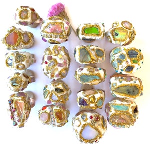 May include: A collection of 18 rings with a white base and gold accents. Each ring is adorned with a variety of colorful stones and crystals, including pink, green, blue, and purple.