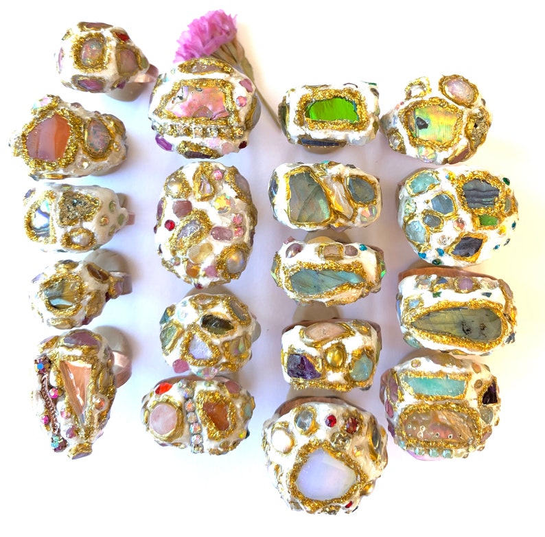 May include: A collection of 18 rings with a white base and gold accents. Each ring is adorned with a variety of colorful stones and crystals, including pink, green, blue, and purple.