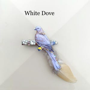 May include: A hair clip featuring a light purple dove figure with iridescent crystal accents and a long, soft beige feather tail. The metal clip base is decorated with small white pearls and clear rhinestones. The text White Dove is visible.