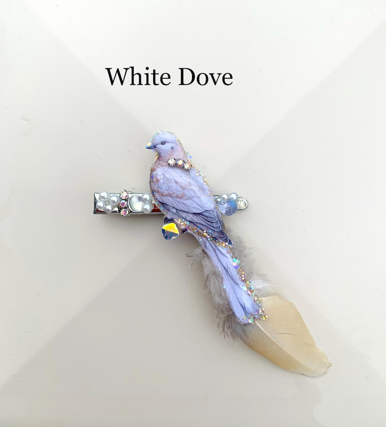 May include: A hair clip featuring a light purple dove figure with iridescent crystal accents and a long, soft beige feather tail. The metal clip base is decorated with small white pearls and clear rhinestones. The text White Dove is visible.