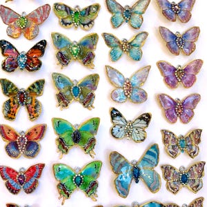 Puede incluir: Una colecci&oacute;n de 24 abalorios de mariposa hechos a mano con detalles intrincados y dise&ntilde;os coloridos. Cada abalorio presenta una mariposa &uacute;nica con diferentes colores, patrones y texturas. Los abalorios est&aacute;n hechos de resina y tienen un acabado dorado.