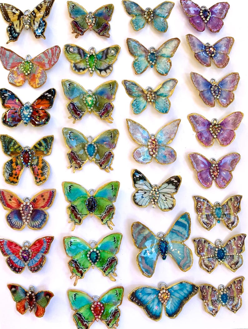 Puede incluir: Una colecci&oacute;n de 24 abalorios de mariposa hechos a mano con detalles intrincados y dise&ntilde;os coloridos. Cada abalorio presenta una mariposa &uacute;nica con diferentes colores, patrones y texturas. Los abalorios est&aacute;n hechos de resina y tienen un acabado dorado.