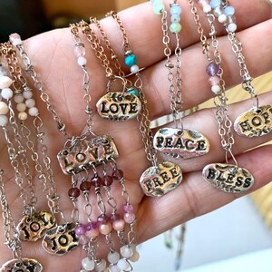 Puede incluir: Una colecci&oacute;n de collares de plata con diferentes dijes y cuentas. Los dijes presentan palabras como "LOVE", "PEACE", "FREE", "BLESS", "HOPE" y "JOY". Las cuentas son en tonos de rosa, blanco y turquesa.