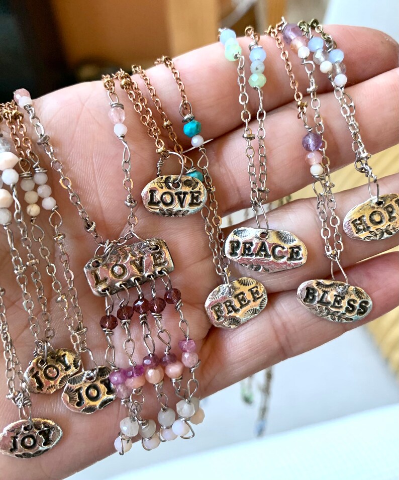 Puede incluir: Una colecci&oacute;n de collares de plata con diferentes dijes y cuentas. Los dijes presentan palabras como "LOVE", "PEACE", "FREE", "BLESS", "HOPE" y "JOY". Las cuentas son en tonos de rosa, blanco y turquesa.