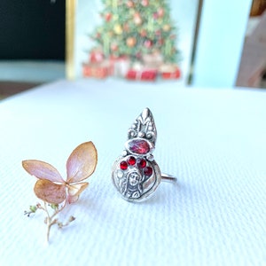 K&ouml;nnte beinhalten: Ein silberner Ring mit einem roten Edelstein und einem detaillierten Design, das ein Frauen Gesicht zeigt. Der Ring liegt auf einer wei&szlig;en Oberfl&auml;che.