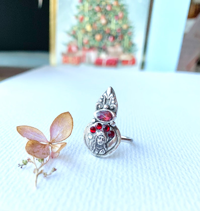 K&ouml;nnte beinhalten: Ein silberner Ring mit einem roten Edelstein und einem detaillierten Design, das ein Frauen Gesicht zeigt. Der Ring liegt auf einer wei&szlig;en Oberfl&auml;che.