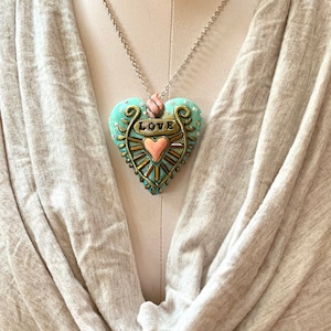 Puede incluir: Un collar con cadena plateada y un colgante en forma de coraz&oacute;n azul turquesa. El coraz&oacute;n tiene detalles dorados y un coraz&oacute;n rosa m&aacute;s peque&ntilde;o en el centro. La palabra "LOVE" est&aacute; escrita en dorado en el colgante.