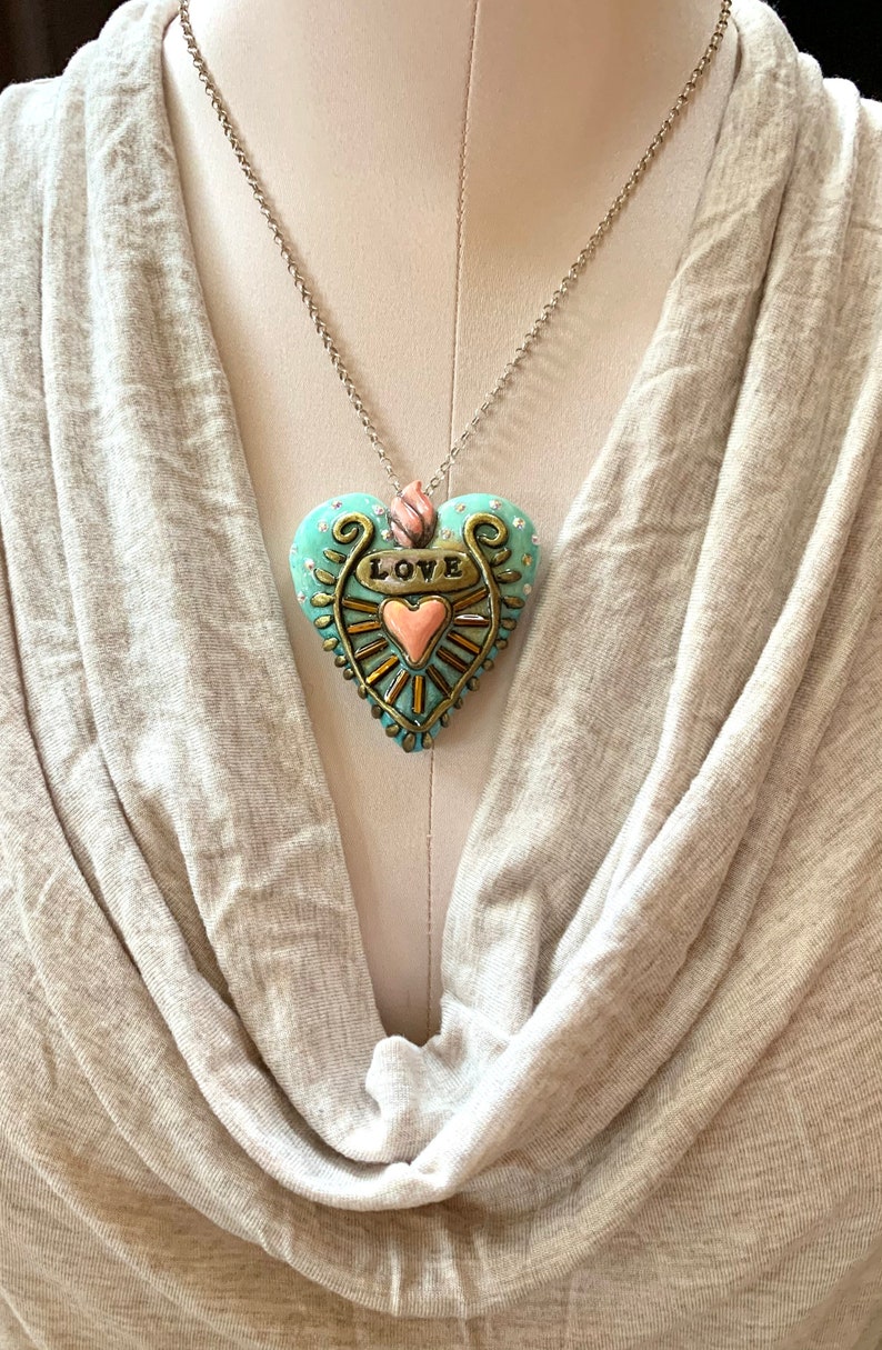Puede incluir: Un collar con cadena plateada y un colgante en forma de coraz&oacute;n azul turquesa. El coraz&oacute;n tiene detalles dorados y un coraz&oacute;n rosa m&aacute;s peque&ntilde;o en el centro. La palabra "LOVE" est&aacute; escrita en dorado en el colgante.