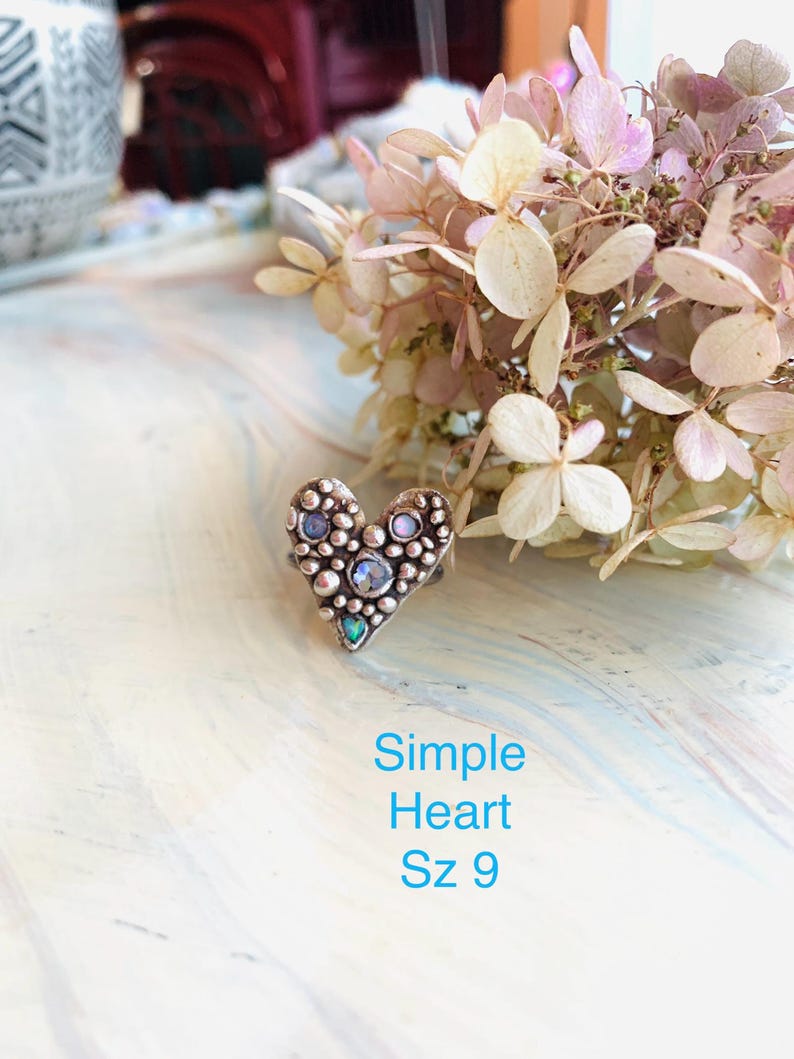 May include: A heart-shaped ring with a textured surface, decorated with small, round embellishments and several iridescent stones. The ring is size 9. The text "Simple Heart Sz 9" is displayed in blue below the ring.