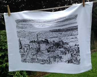 May include: A white tea towel with a black and white ink drawing of a city skyline. The drawing features a castle and many buildings.