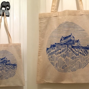 Puede incluir: Dos bolsas de lona con diseños impresos en azul del Castillo de Edimburgo. La bolsa más grande está colgada de una puerta blanca, mientras que la bolsa más pequeña está colgada de un gancho.