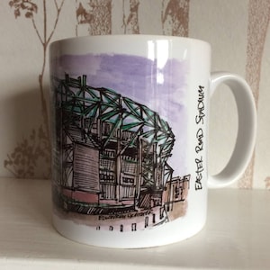 May include: White ceramic mug featuring a watercolour illustration of a stadium exterior. The stadium is green and brown with a purple sky in the background. The text "Easter Road Stadium" is printed on the side of the mug.