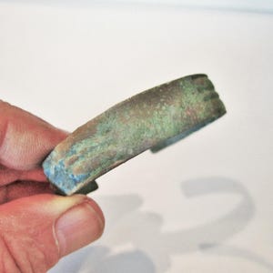 Könnte beinhalten: Ein Armreif mit einer verwitterten, oxidierten Oberfläche in Grün-, Blau- und Brauntönen. Das Metallband hat eine dicke, massive Konstruktion mit einem leicht gewölbten Profil und einem offenen Design an der Rückseite mit texturierter Optik.