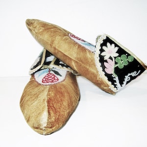 Puede incluir: Un par de mocasines hechos a mano con exterior de cuero marrón claro. Las partes superiores están adornadas con intrincados abalorios con diseños florales en rosa, verde y blanco sobre un fondo negro. Los mocasines tienen una estética tradicional nativa americana.