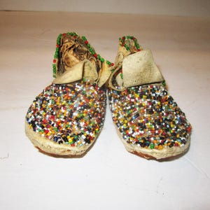 May include: A pair of beaded moccasins with colourful beads in shades of red, green, blue, white, and black. The moccasins are made of leather and have a fringe detail.