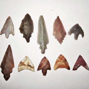 May include: A collection of ten ancient arrowheads in various shapes and sizes. The arrowheads display a range of colors, including tan, brown, gray, and reddish hues. They are likely made of stone or flint, and are arranged on a white surface.