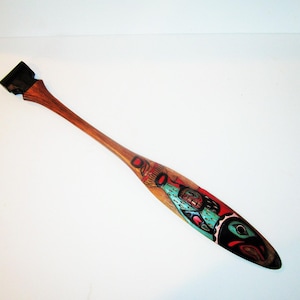 May include: A wooden paddle with a dark handle and a blade featuring a colorful design. The blade has red, black, and teal accents, with a carved design of a face and other shapes. The paddle is approximately 40 inches long.