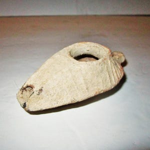 May include: A beige, teardrop-shaped oil lamp made of textured, porous material. The lamp features a central circular opening for oil and a small nozzle at the pointed end, with subtle vertical ridges along the curved sides of the body.