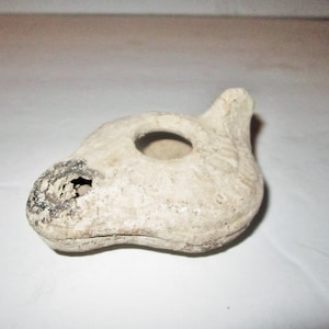 May include: An off-white, textured ceramic oil lamp with a central circular opening and a protruding nozzle. The surface features faint, molded decorative patterns and a darkened, charred area around the wick hole at the front.