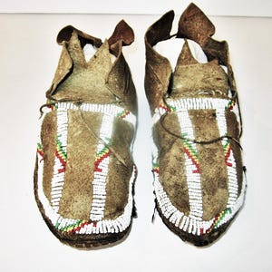 May include: A pair of brown leather moccasins featuring intricate beadwork in white, red, yellow, and green. The footwear has a soft, flexible construction with a gathered toe seam and a high heel collar, showcasing detailed geometric patterns.
