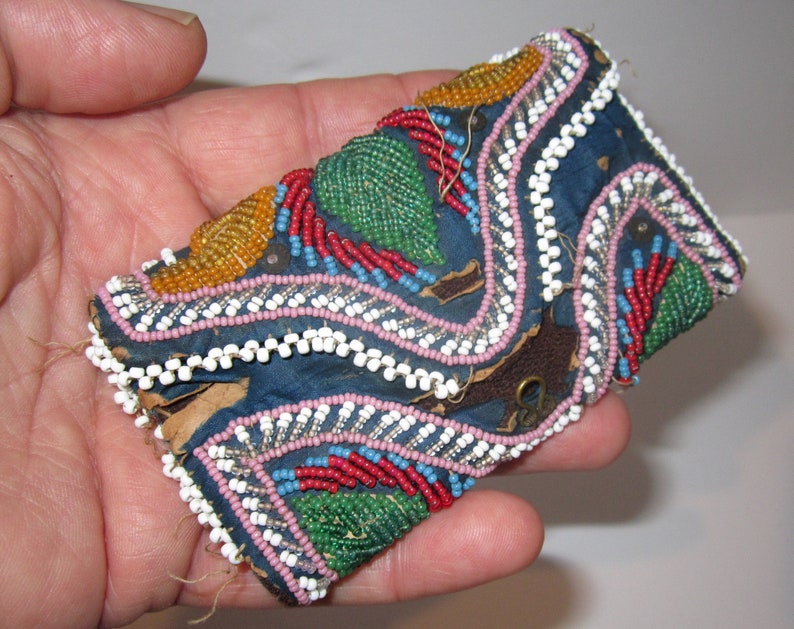 May include: A small, blue fabric pouch with a flap closure, decorated with colorful beads in a geometric pattern. The pouch has a metal clasp and is hand-stitched.