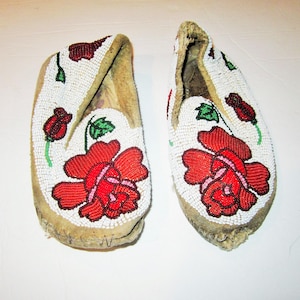 May include: A pair of beaded moccasins. The white surface is adorned with red roses, buds, and green leaves. The edges are trimmed with white beads. The soles are made of a light brown material.
