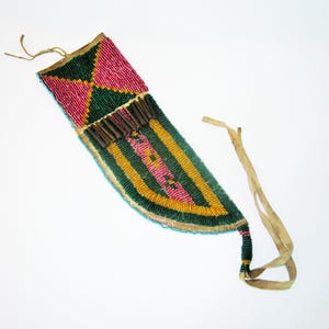 May include: A beaded knife sheath with a colorful design. The top section features a pink and green geometric pattern. The main body has vertical stripes in yellow, pink, and green. A leather strap extends from the bottom.