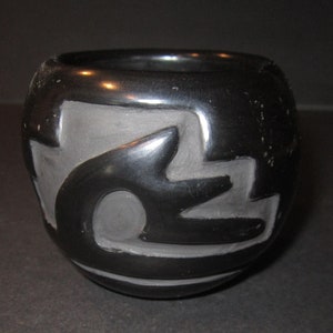 May include: Black pottery bowl with a carved design. The design is a stylized geometric pattern with a central circle.
