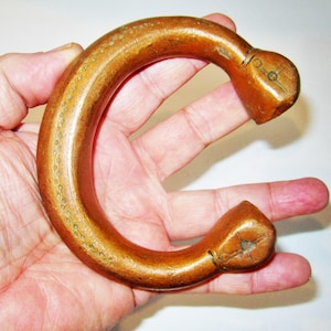 May include: A curved, wooden bracelet with a warm, brown hue. The bracelet has a rounded shape with decorative details and is held in a person's hand. The ends of the bracelet have a squared-off design. The bracelet is approximately 3 inches in diameter.