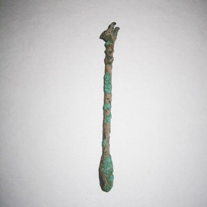 Può includere: Un'asta metallica lunga e sottile con un'estremità appuntita e una testa arrotondata. L'asta è ricoperta di patina verde.